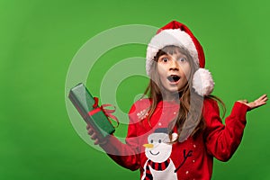 Portrait of an emotional surprised girl in a red knitted sweater and a Santa hat holds a beautiful gift in her hand
