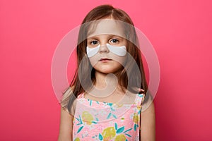 Portrait of emotional upset female child posing isolated over rose background, has white patches under eyes, standing with serious