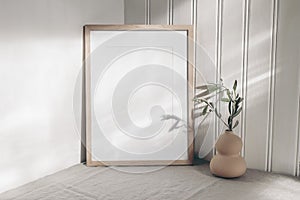 Portrait empty wooden frame mockup in sunlight. Olive branch in modern organic shaped vase. Beige linen table cloth