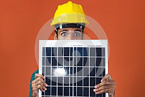 Portrait of an engineer or solar worker in protective work clothes holding photovoltaic panel isolated on orange background