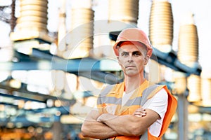 Portrait of engineer worker in uniform and helmet standing near high voltage substation with tall pylons and voltage
