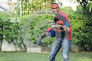 Portrait of enjoy African American father carrying little cute son flying in superhero costume in garden at home. Happy loving