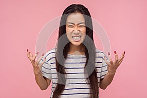 Portrait of enraged furious girl with long hair in striped t-shirt standing with clenched teeth and raised hands