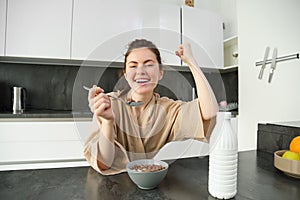 Portrait of enthusiastic young woman eating cereals with milk, looking excited and happy, sitting near kitchen worktop