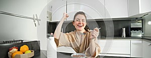 Portrait of enthusiastic young woman eating cereals with milk, looking excited and happy, sitting near kitchen worktop
