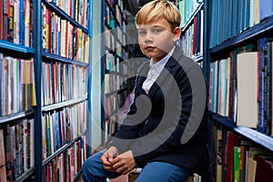 portrait of erudite boy sitting among books in library