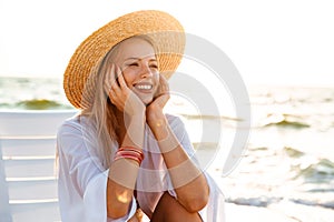 Portrait of european cheerful woman 20s in straw hat smiling, while sitting in lounge chair at seaside during summer morning