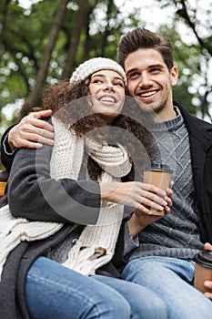 Portrait of european people man and woman 20s drinking takeaway coffee from paper cups, while sitting on bench in green park