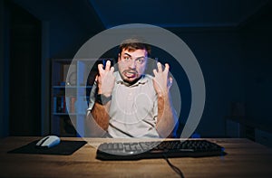 Portrait of an evil man in a white T-shirt at night on the computer looks at the camera and shows anger. Freelancer is nervous in