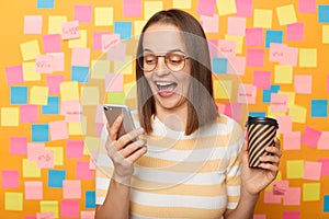 Portrait of excited amazed happy woman wearing striped T-shirt posing over sticky notes to write reminder on yellow wall, drinking