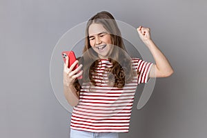 Amazed little girl standing holding phone, looking at camera and celebrate victory with fist.