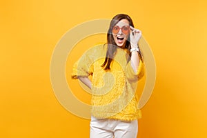 Portrait of excited amazed young woman in fur sweater, white pants holding heart orange eyeglasses isolated on bright