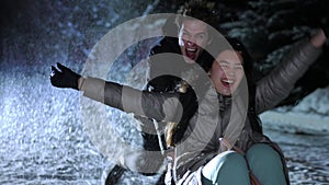 Portrait of excited Asian girl laughing posing as Caucasian boy pushing sled riding on winter night outdoors. Happy