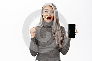 Portrait of excited asian senior woman showing mobile phone screen and celebrating, achieve goal on smartphone app