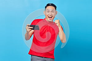 Portrait of excited asian young man playing game on mobile phone and doing celebration gesture for winning game isolated on blue