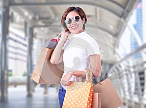Portrait of an excited beautiful asian girl wearing dress and sunglasses holding shopping bags while standing at fashion mall