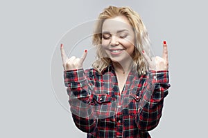 Portrait of excited beautiful blonde young woman in casual red checkered shirt standing with closed eyes, toothy smile rock sign