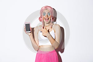 Portrait of excited beautiful girl in halloween costume, pink wig and bright makeup, pointing finger at mobile phone
