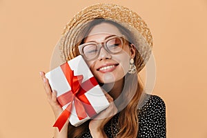 Portrait of excited beautiful woman 20s wearing straw hat and sunglasses smiling while holding gift box with red ribbon, isolated