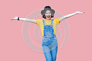 Portrait of excited beautiful young woman in yellow t-shirt, blue denim overalls, makeup, black hat standing with raised arms and