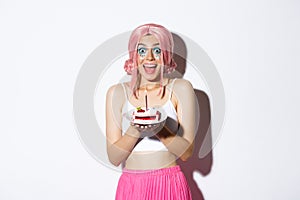 Portrait of excited birthday girl in pink wig, looking amused while making wish on b-day cake with lit candle