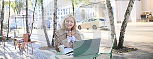 Portrait of excited blond woman, student in outdoor cafe, sitting in coffee shop with laptop, looking happy and amused
