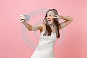 Portrait of excited bride woman in lace white wedding dress showing victory sign, doing taking selfie shot on mobile