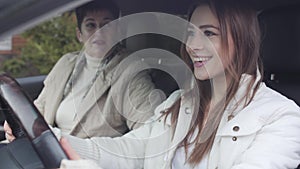 Portrait of excited Caucasian girl sitting on driver`s seat and listening to wise woman explaining how to drive. Happy