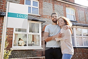 Portrait Of Excited Couple Standing Outside New Home With Sold Sign