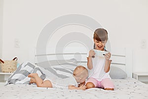Portrait of excited female child wearing white t shirt and pink shorts playing video games on cellphone, cute infant baby lying on