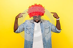 Portrait of excited funny carefree man pointing at red curly wig on his head.  on yellow background