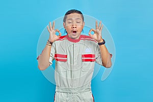 Portrait of excited handsome man wearing mechanic uniform showing okay gesture with open mouth over blue background