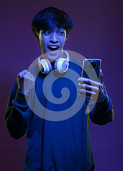 Portrait of excited handsome young asian man looking at the mobile phone wearing headphone in neon light