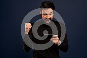 Portrait of excited, happy bearded man holding mobile phone horizontally and smiling amused at display, playing awesome new game,