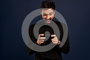 Portrait of excited, happy bearded man holding mobile phone horizontally and smiling amused at display, playing awesome new game,