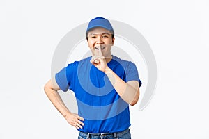 Portrait of excited happy courier making surpirse. Delivery guy in blue uniform asking keep quiet, smiling and shushing