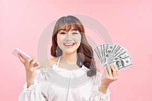 Portrait of an excited happy girl showing bunch of money banknotes while holding mobile phone and looking at camera isolated over