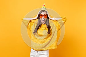 Portrait of excited joyful young woman in orange heart glasses and birthday party hat screaming, putting hands on head