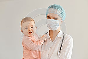 Portrait of excited kid and young adult female pediatrist wearing medical uniform, woman holding infant baby girl, examining child