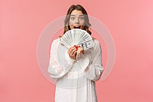 Portrait of excited lucky girl with blond hair, white dress, winning money, receive cash award, big lottery prize