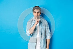 Portrait of excited man asking to keep quiet, showing hush taboo sign and looking nervously at camera, standing against