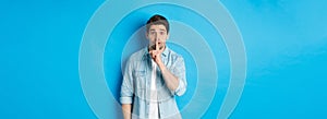 Portrait of excited man asking to keep quiet, showing hush taboo sign and looking nervously at camera, standing against