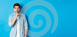 Portrait of excited man asking to keep quiet, showing hush taboo sign and looking nervously at camera, standing against