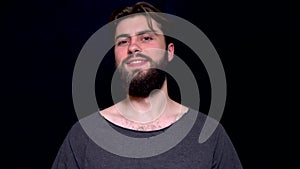 Portrait of excited man with opened mouth giving thumbs-up and winking against black background. Smiling bearded young