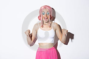 Portrait of excited party girl in pink wig and bright makeup, react to something amazing, pointing fingers down and