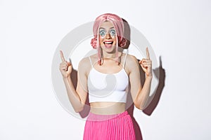 Portrait of excited party girl with pink wig and halloween makeup, looking amazed while pointing fingers up, standing
