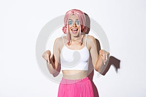 Portrait of excited party girl in pink wig and halloween outfit, looking amused and happy while pointing fingers down