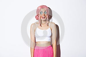 Portrait of excited party girl showing tongue and smiling, looking left with happy expression, wearing pink wig for