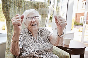 Portrait Of Excited Senior Woman Sitting In Chair In Lounge Of Retirement Home