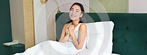 Portrait of excited, smiling asian woman sitting in her bed in morning, daydreaming, looking hopeful